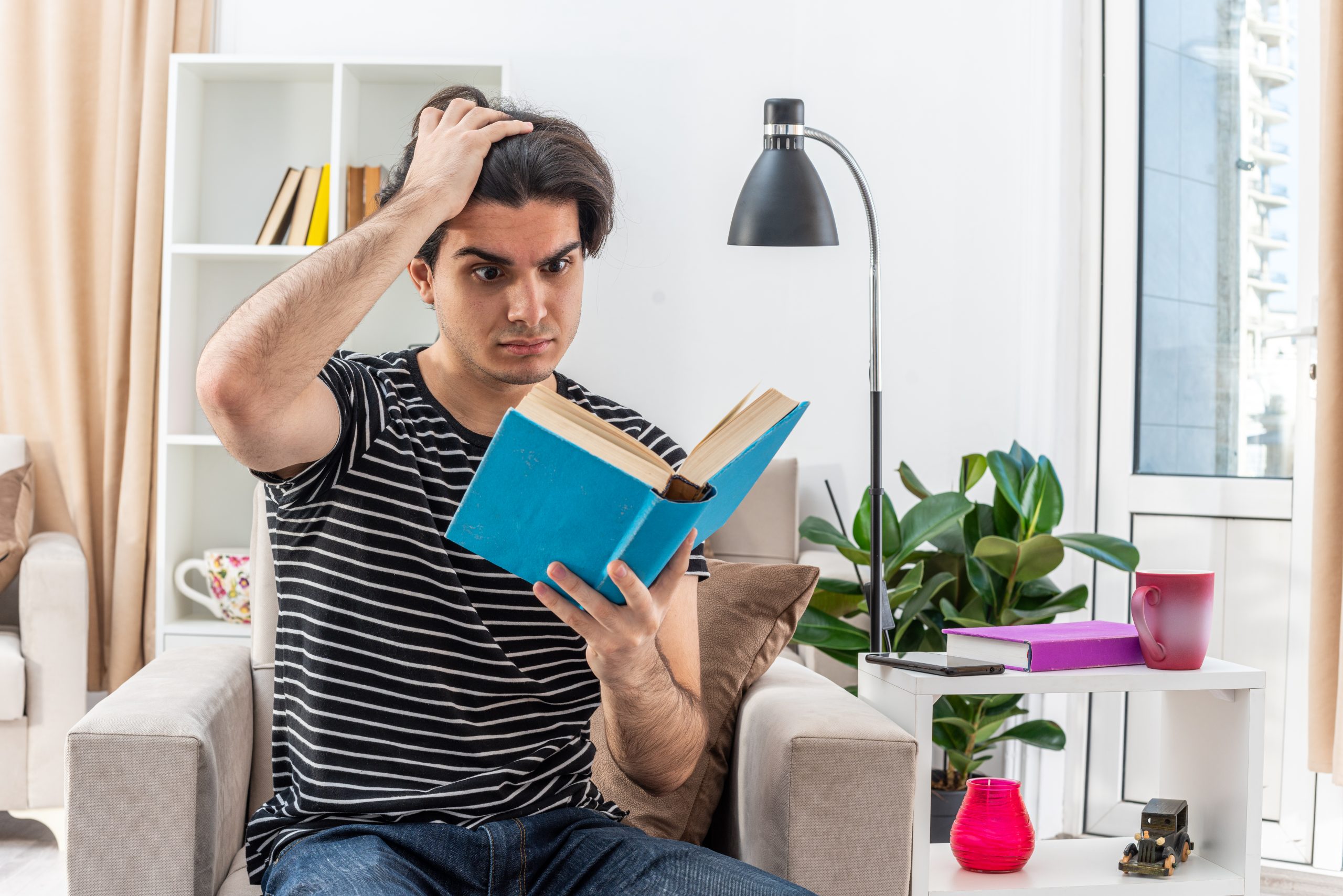young man in casual clothes hlding book reading looking puzzled sitting on the chair in light living room
